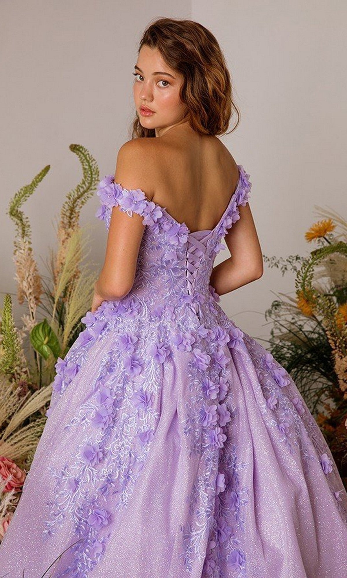 A young woman in the Eureka 9339 long prom dress, a lavender off-shoulder glitter ball gown with 3D floral appliques, stands looking over her shoulder, surrounded by decorative plants.